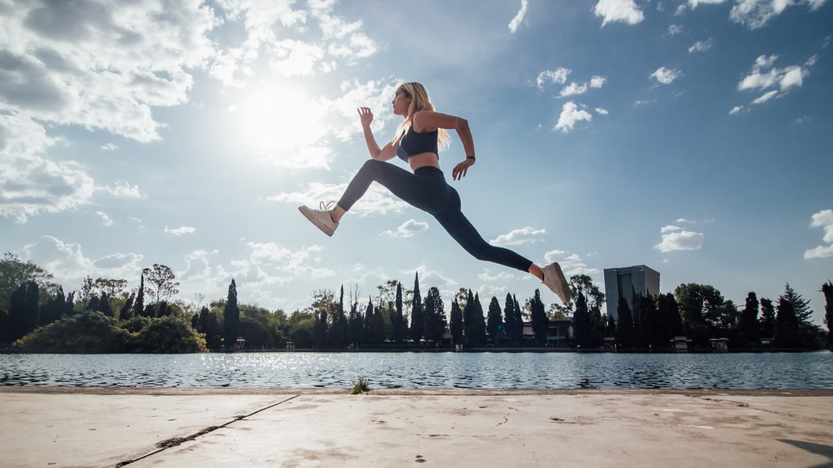 Woman performs an athletic jump by a lakeside in the park under a sunny blue sky.